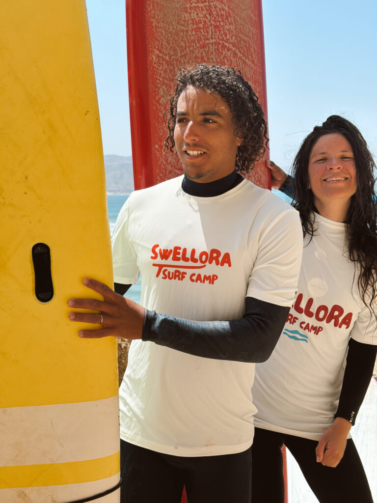 Surf School Imsouane Instructors Yassine and Camille Two smiling surf coaches in Swellora Surf Camp shirts holding a yellow soft top surfboard in Imsouane